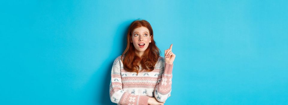 Thoughtful redhead girl in winter sweater having an idea, raising finger in eureka sign and looking up, standing against blue background.