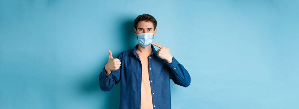 Coronavirus and pandemic concept. Young healthy man pointing at medical mask and showing thumbs up, using preventive measures from catching covid-19, blue background.