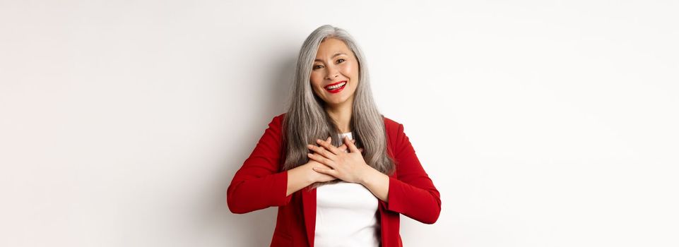 Business concept. Mature asian woman with red lips and blazer, holding hands on heart and smiling thankful, looking grateful at camera, standing over white background.