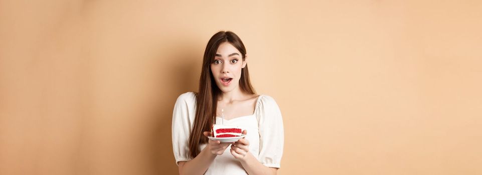 Excited birthday girl looking amazed, holding cake with candle, making b-day wish, standing on beige background.