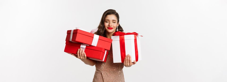 Holidays, celebration concept. Beautiful caucasian woman in elegant dress holding Christmas presents and smiling happy, standing over white background.