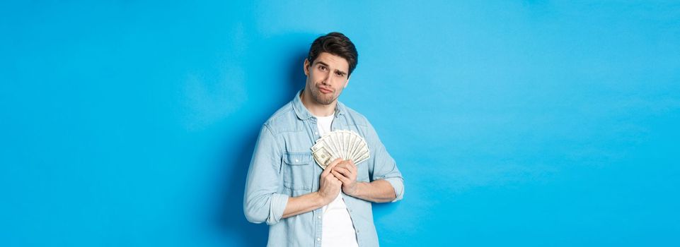 Handsome young man keeping money to himself, smiling and looking greedy, standing over blue background.