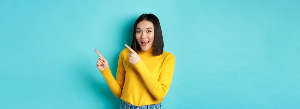 Shopping concept. Beautiful chinese girl in yellow sweater pointing fingers at upper right corner logo banner, smiling amused, standing over blue background.