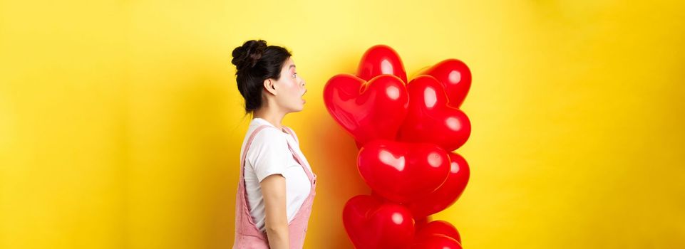 Valentines day and relationship concept. Profile of surprised asian girl staring left at logo, standing near heart balloons, yellow background.