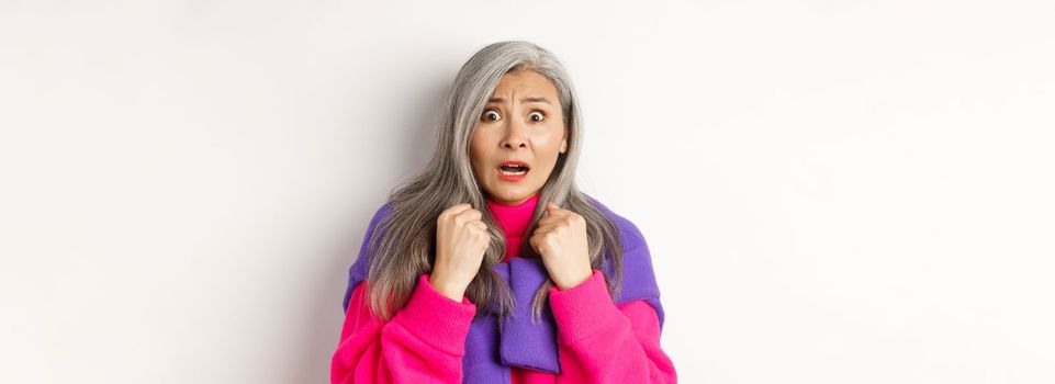 Close up of scared asian senior woman gasping, looking frightened and startled at camera, standing over white background.