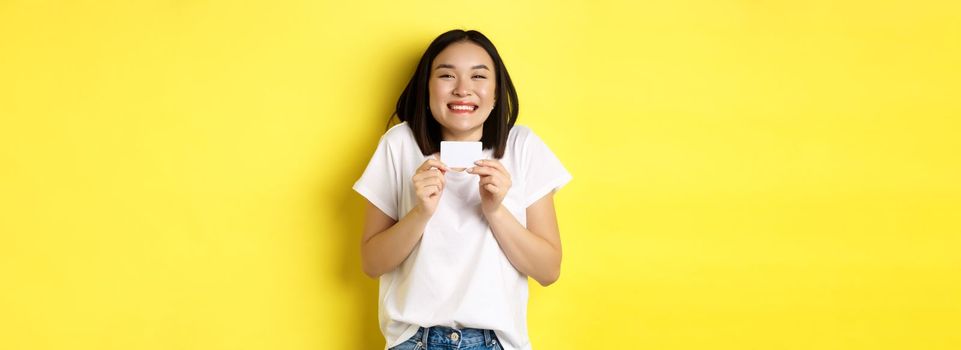 Young asian woman in casual white t-shirt going on shopping, showing plastic credit card and smiling excited, standing over yellow background.