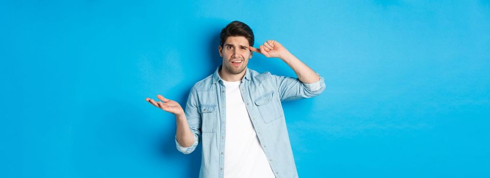 Confused man scolding person for being stupid, pointing finger at head and shrugging, looking puzzled, disapprove actions, standing against blue background.