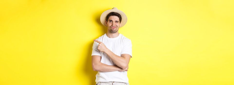 Skeptical male tourist complaining, pointing finger right at something bad or lame, standing unamused against yellow background.