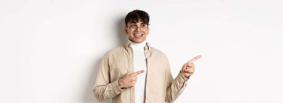 Excited and enthusiastic young man in glasses, pointing fingers and looking left with happy smile, standing in awe on white background.