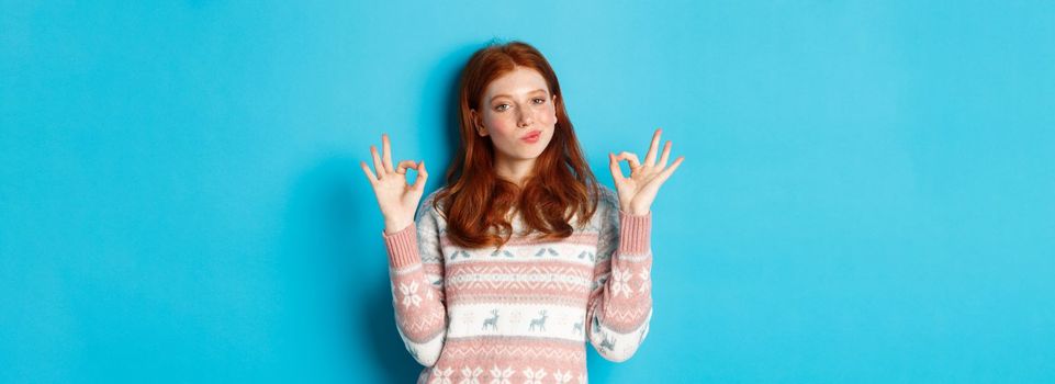 Satisfied and proud redhead girl nod in approval, showing okay sign, not bad or praise gesture, standing against blue background.