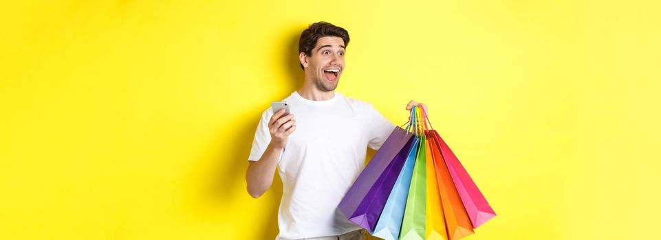 Concept of mobile banking and cashback. Happy man looking amazed, holding shopping bags and smartphone, yellow background.