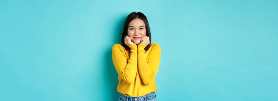 Beauty and fashion concept. Beautiful asian woman blushing and smiling, looking at something cute and silly, lean face on palms, standing over blue background.