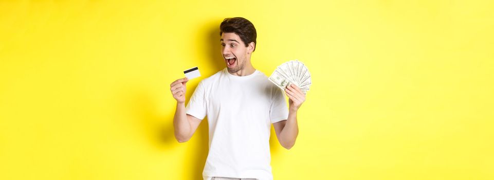 Cheerful guy looking at credit card, holding money, concept of bank credit and loans, standing over yellow background.