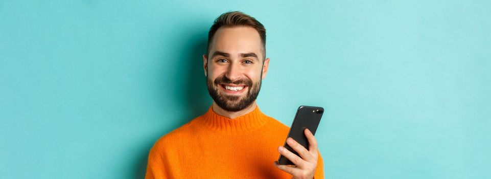 Close-up of happy handsome man writing message on mobile phone, holding smartphone and smiling, standing against turquoise background.
