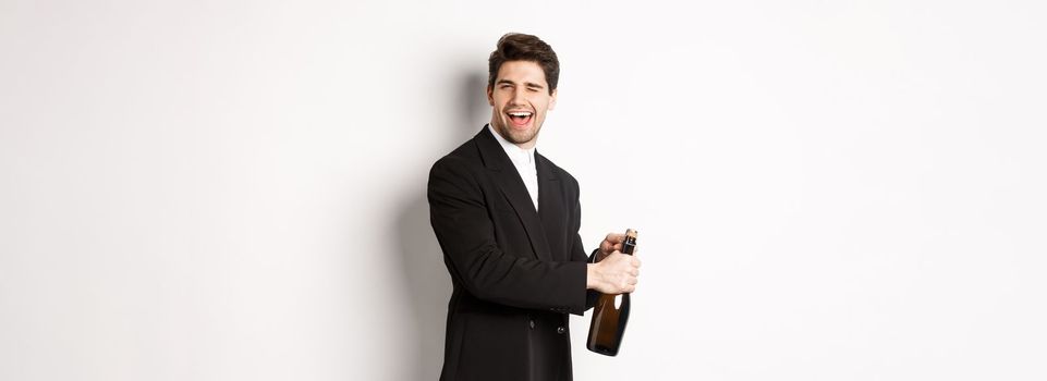 Portrait of attractive man in black suit, winking at camera and opening bottle of champagne, celebrating new year, standing against white background.