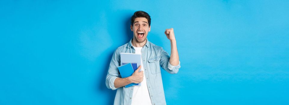 Cheerful guy holding notebooks and celebrating, making fist pump and shouting yes with excitement, standing over blue background.