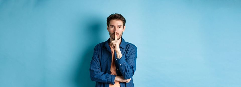 Modern guy shushing at camera, press finger to lips and telling a secret, making taboo sign, standing on blue background.