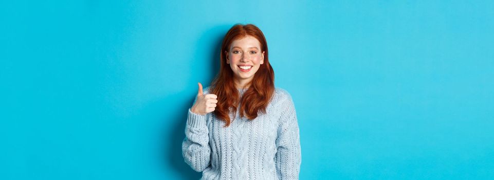 Cute redhead girl in sweater showing thumb up, like and agree, smiling pleased, standing against blue background.
