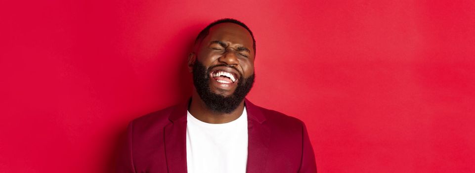 Close-up of happy and carefree Black man having fun, laughing and smiling, standing in blazer against red background.