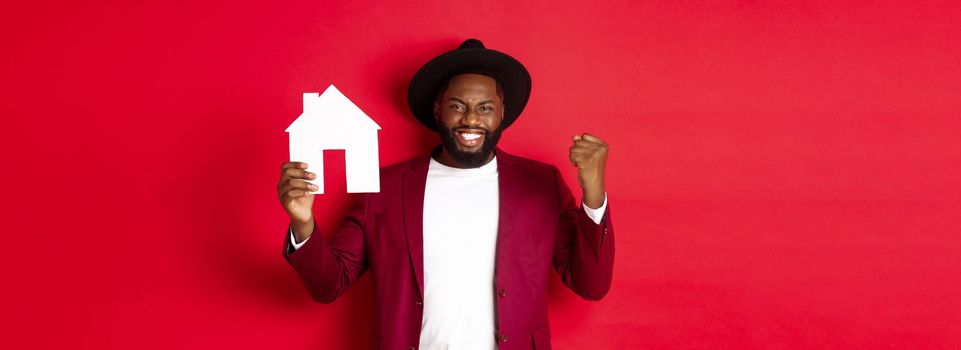 Real estate. Cheerful Black man rejoicing and showing paper home maket, standing over red background.