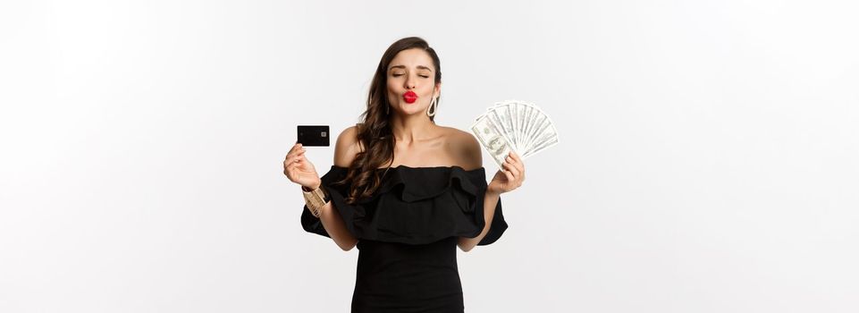 Beauty and shopping concept. Pretty glamour woman pucker lips for kiss, showing credit card and dollars, standing over white background.