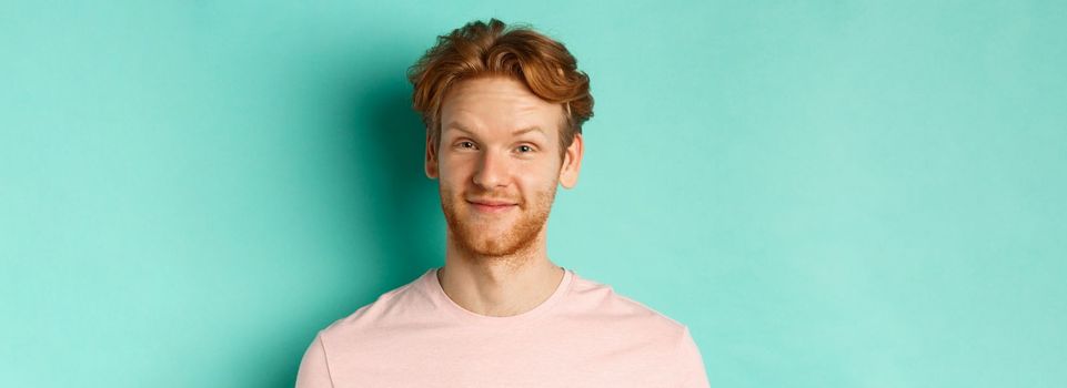 Close up of redhead bearded man looking pleased, nod in approval and smiling, standing in pink t-shirt against turquoise background.