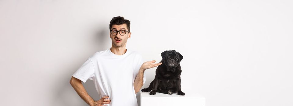 Confused man seeing something strange, staring at camera puzzled, standing near cute black pug pet against white background.