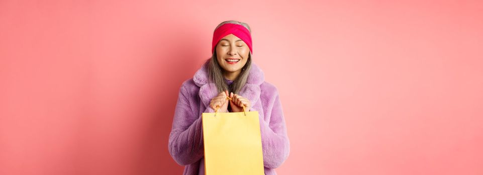 Shopping and fashion concept. Happy asian senior woman in stylish clothes looking happy and delighted, standing with yellow paper bag and smiling, pink background.