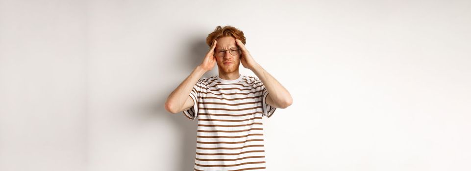 Image of young man with red hair and glasses touching head, frowning from painful migraine, having headache, standing over white background.