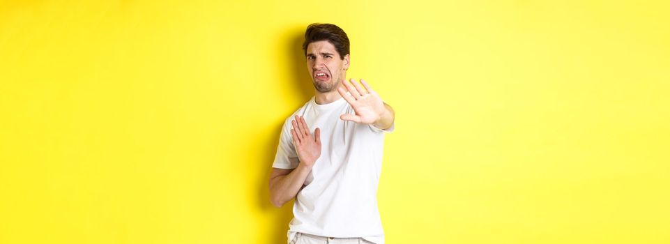 Disgusted man refusing, grimacing from dislike and aversion, begging to stop, standing in white t-shirt against yellow background.