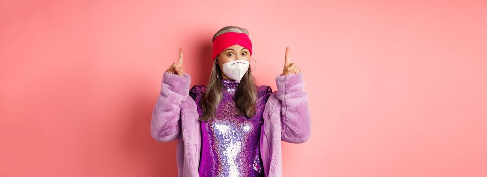 Covid and fashion concept. Fashionable asian woman in glitter dress and face mask, pointing fingers up, showing advertisement, pink background.