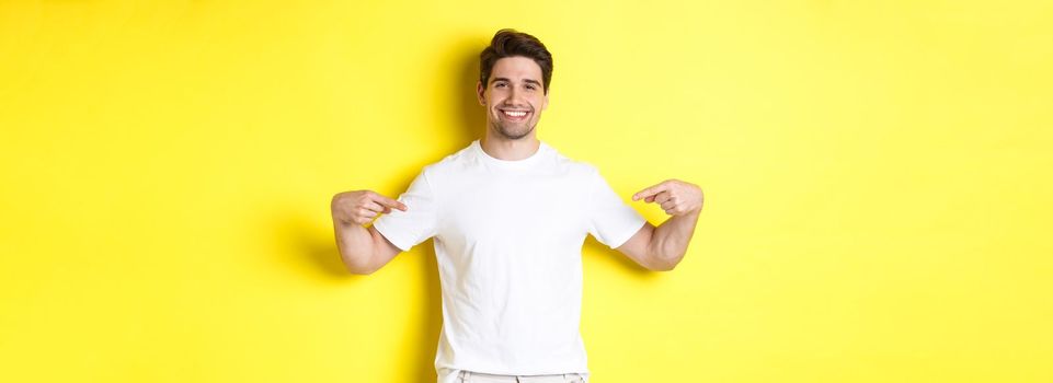 Happy attractive guy pointing fingers at your logo, showing promo on his t-shirt, standing over yellow background.