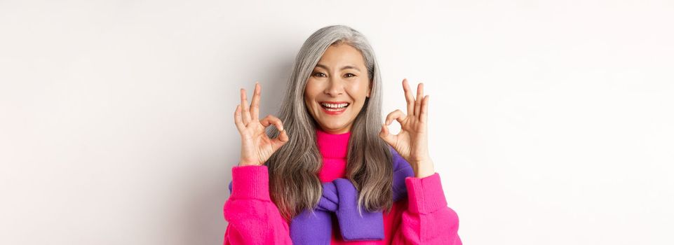 Close-up of satisfied asian female with grey hair, smiling pleased and showing OK signs, approve and like product, praising something awesome, white background.