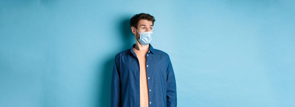 Covid-19 and healthcare concept. Surprised caucasian guy in face mask looking aside at empty space for banner, standing on blue background.