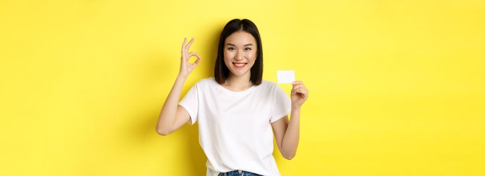 Young asian woman in casual white t-shirt showing plastic credit card and okay gesture, recommend bank, smiling at camera, yellow background.