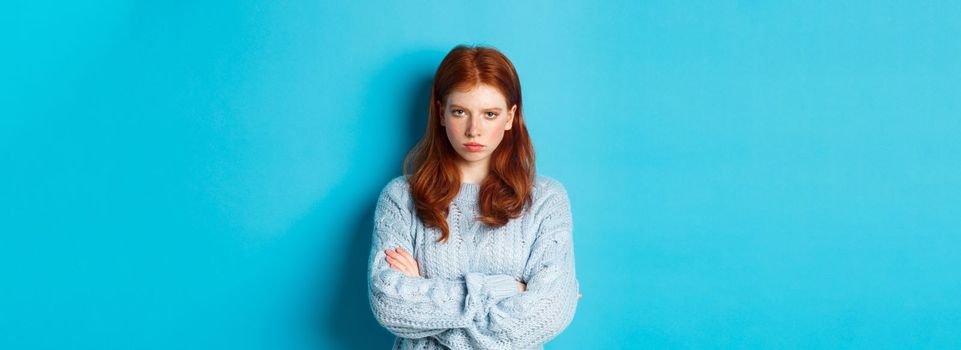 Image of angry redhead girl feeling offended, cross arms on chest and sulking, staring at camera mad, standing against blue background.