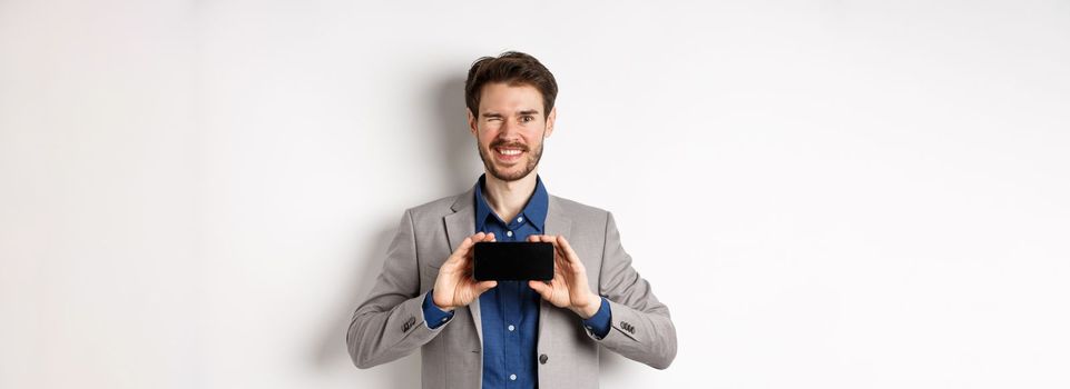 E-commerce and online shopping concept. Cheeky male model in suit winking and showing empty smartphone screen, smiling happy at camera, white background.