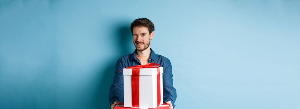 Handsome boyfriend winking at camera and wishing happy valentines day, extending hands with gift boxes, making romantic surprise, standing over blue background.