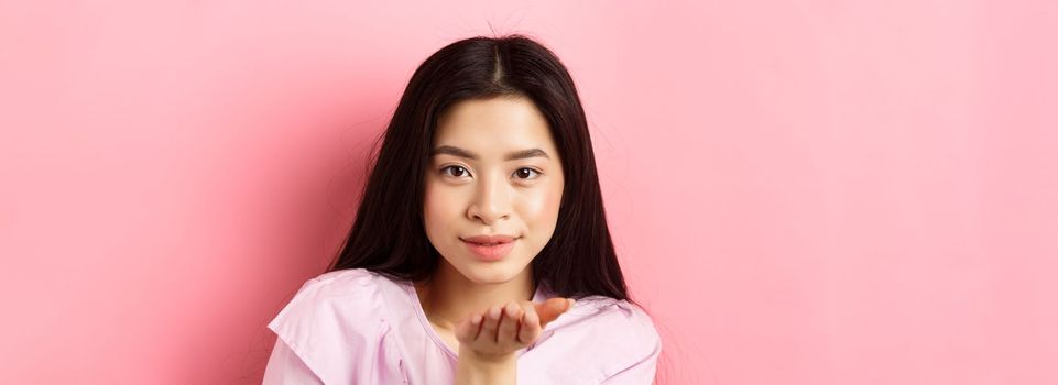 Beautiful asian girl with natural makeup, blowing air kiss at camera with sensual gaze, standing romantic against pink background.