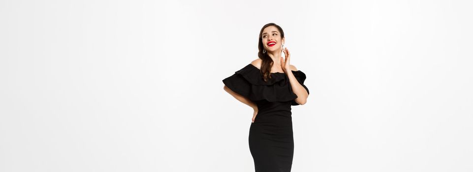 Celebration and christmas holidays concept. Beautiful woman in black dress with gifts and looking surprised, standing over white background.