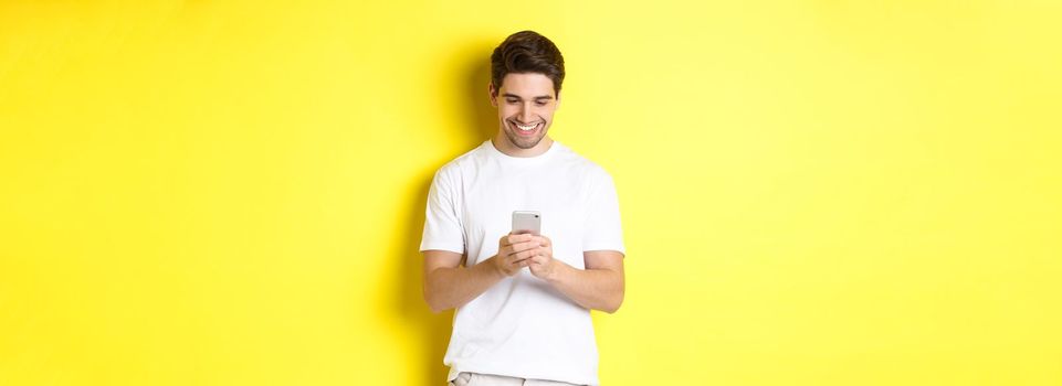 Young man reading text message on smartphone, looking at mobile phone screen and smiling, standing in white t-shirt against yellow background.