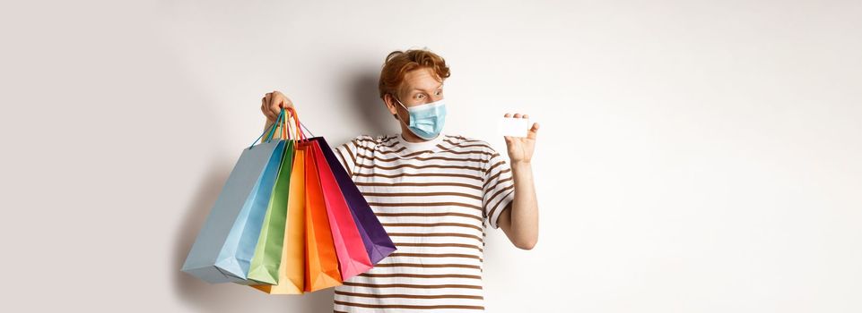 Pandemic and special offer concept. Happy redhead man in face mask showing shopping bags and plastic credit card, standing over white background.