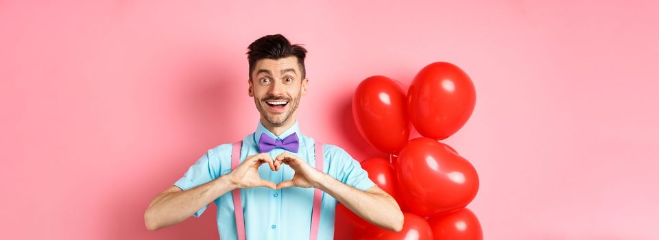 Valentines day concept. Romantic guy looking happy and smiling, showing heart gesture to lover on special anniversary, standing on pink background.