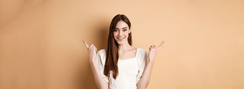 Beautiful young woman in dress pointing fingers sideways, show two ways, giving choice and smiling, standing on beige background.