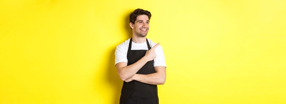 Handsome barista pointing and looking left at promo, wearing black apron, standing against yellow background.