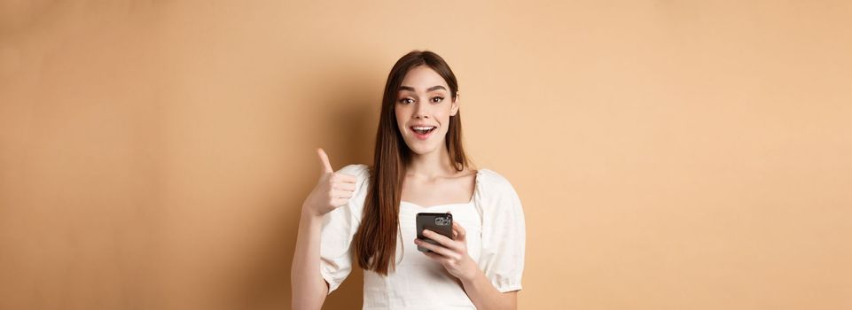 Online shopping concept. Happy young woman showing thumb up and using cellphone, smiling pleased, standing on beige background.