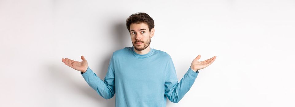 Indecisive young man with beard, shrugging shoulders and know nothing, looking away confused, standing in blue shirt over white background.