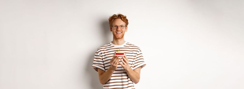 Holidays and celebration concept. Handsome redhead man in glasses holding birthday cake with candle, smiling and looking happy at camera.