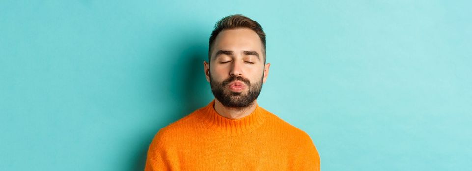 Close-up of young man in sweater pucker lips, close eyes and waiting for kiss, standing over light blue background.