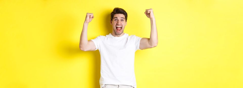 Image of excited man winning, raising hands up and celebrating, triumphing and rooting for team, standing over yellow background. Copy space